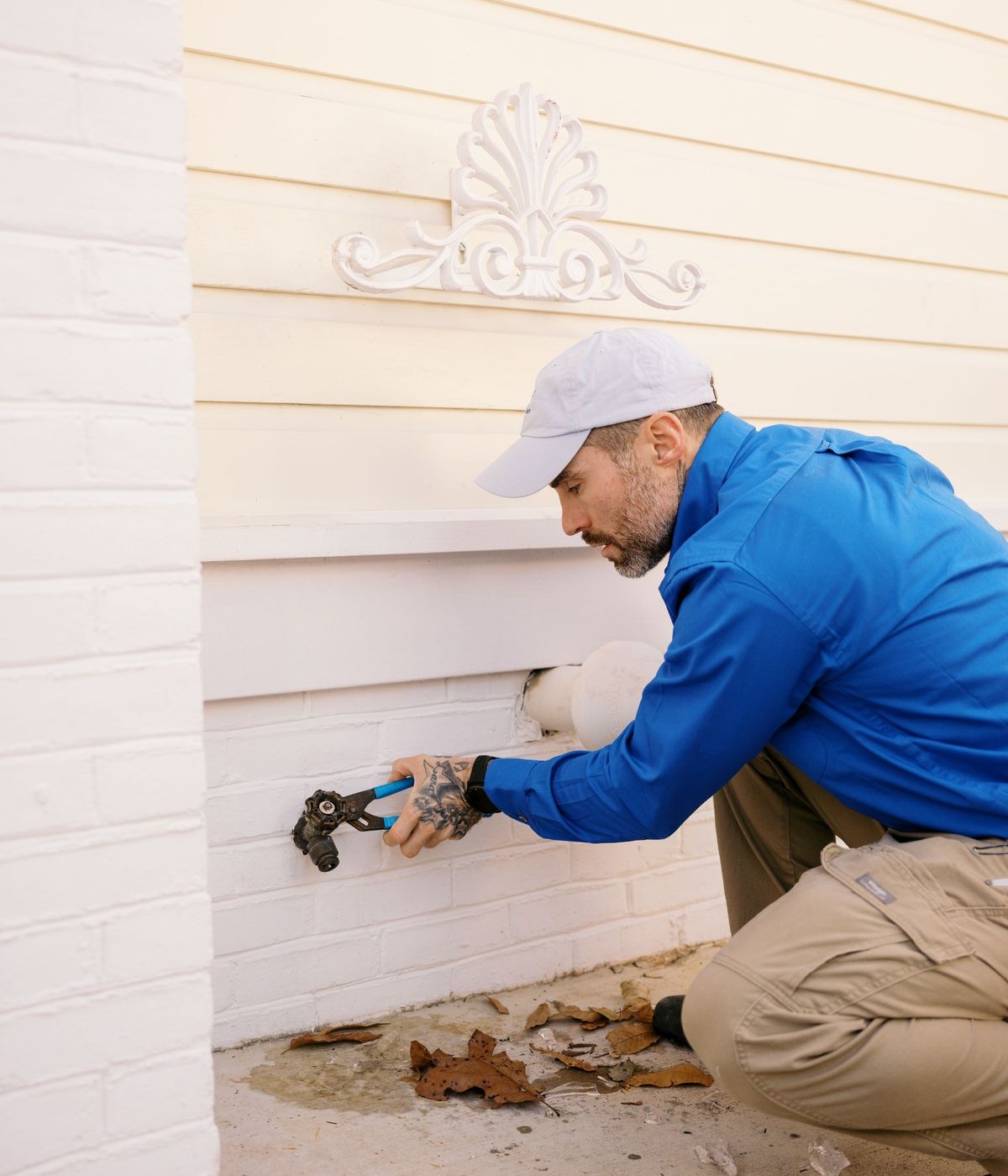 Plumber In Blue Uniform Repairing Outdoor Water Spigot Leak On White Brick Wall
